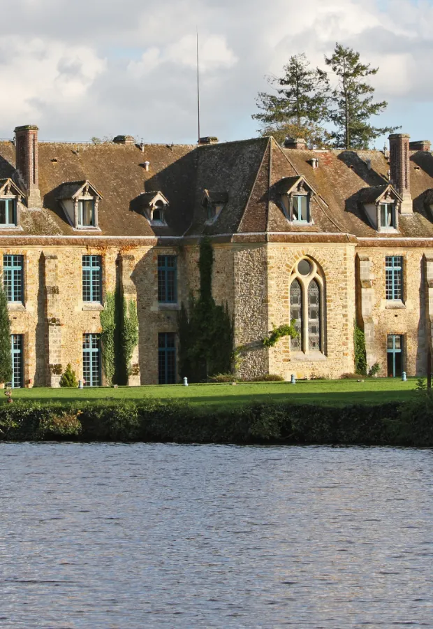 Stone historic building with blue windows and steep roof beside a body of water under a cloudy sky.