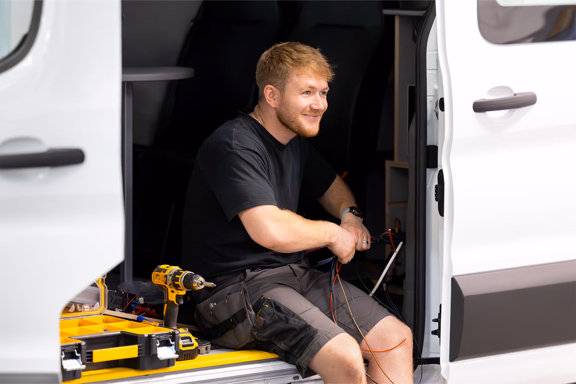 Young fitter holding holding cables sat in the doorway of a panel van. Drill and tools to his side.