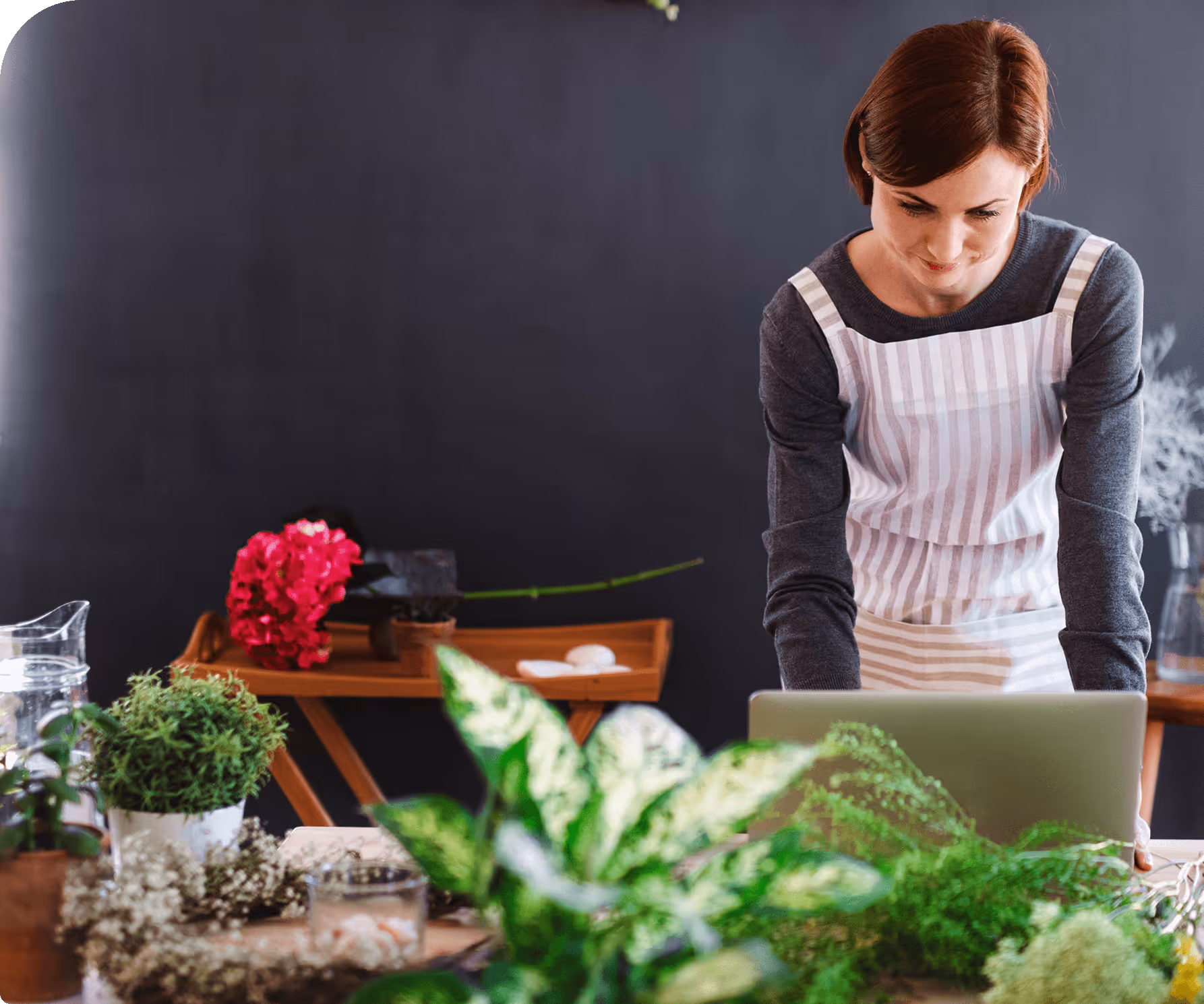 Woman in apron working on a laptop behind a table with various green plants and flowers.