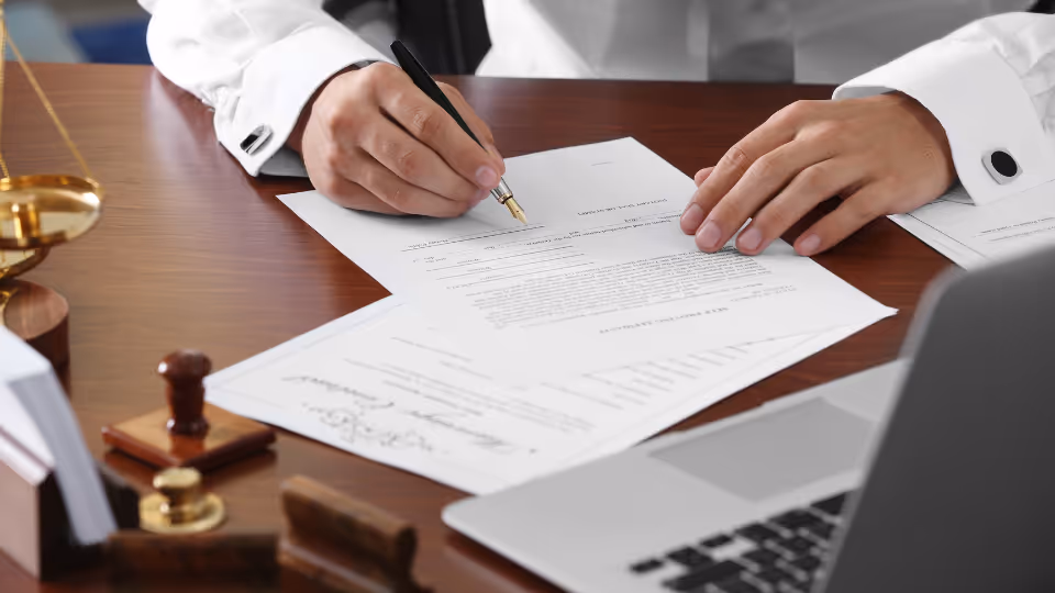 Alt text: A person signing a document on a desk with a laptop