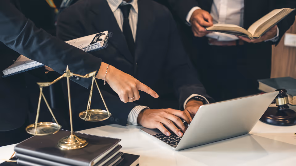 Alt text: Three lawyers in suits sitting at a desk with a laptop and a scale of justice