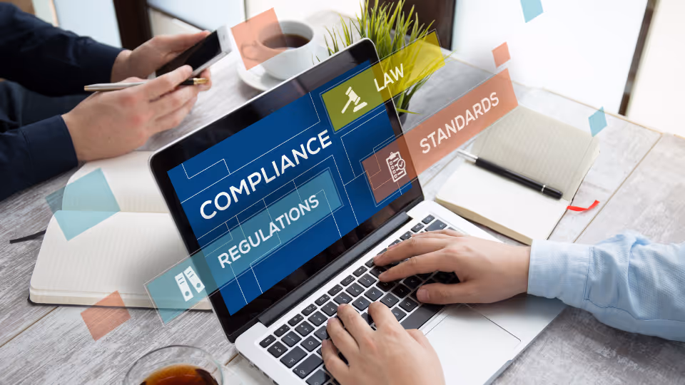 Alt text:  A laptop on a desk with the words "Compliance," "Law," "Regulations," and "Standards" floating above the screen in bold text