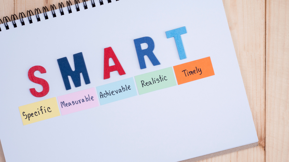 Alt text: A notebook open on a wooden table with the acronym "SMART" written on it, along with its meanings: Specific, Measurable, Achievable, Relevant, and Time-bound