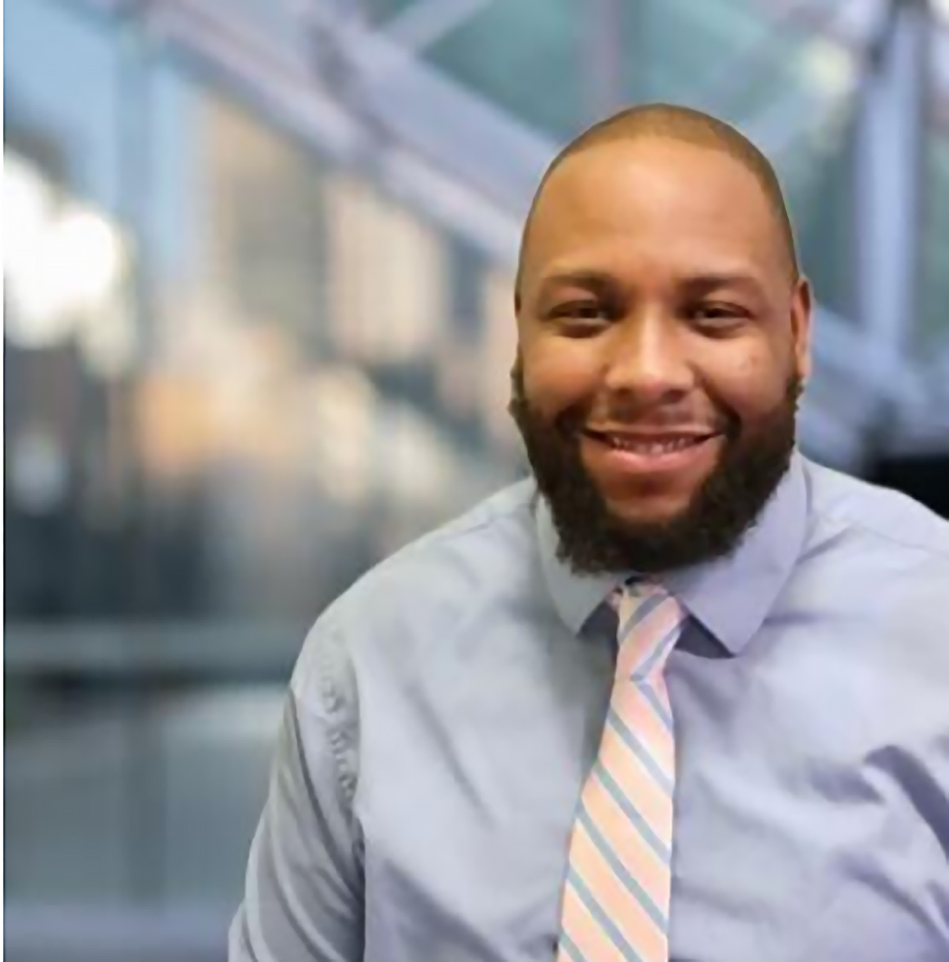 Smiling man with a beard wearing a light blue shirt and striped tie in an office setting.