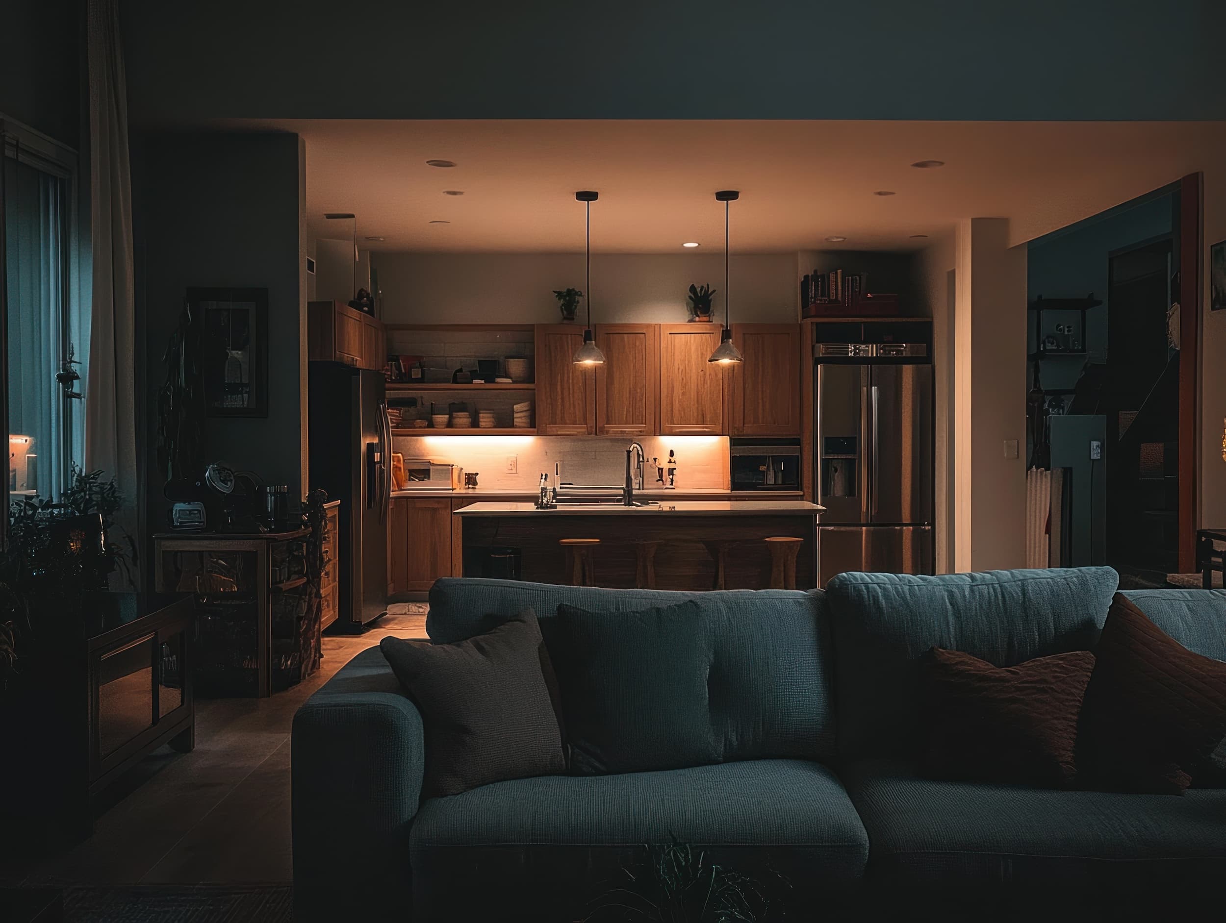 Cozy modern kitchen with wooden cabinets, pendant lights, and a dark blue sofa in the foreground at dusk.