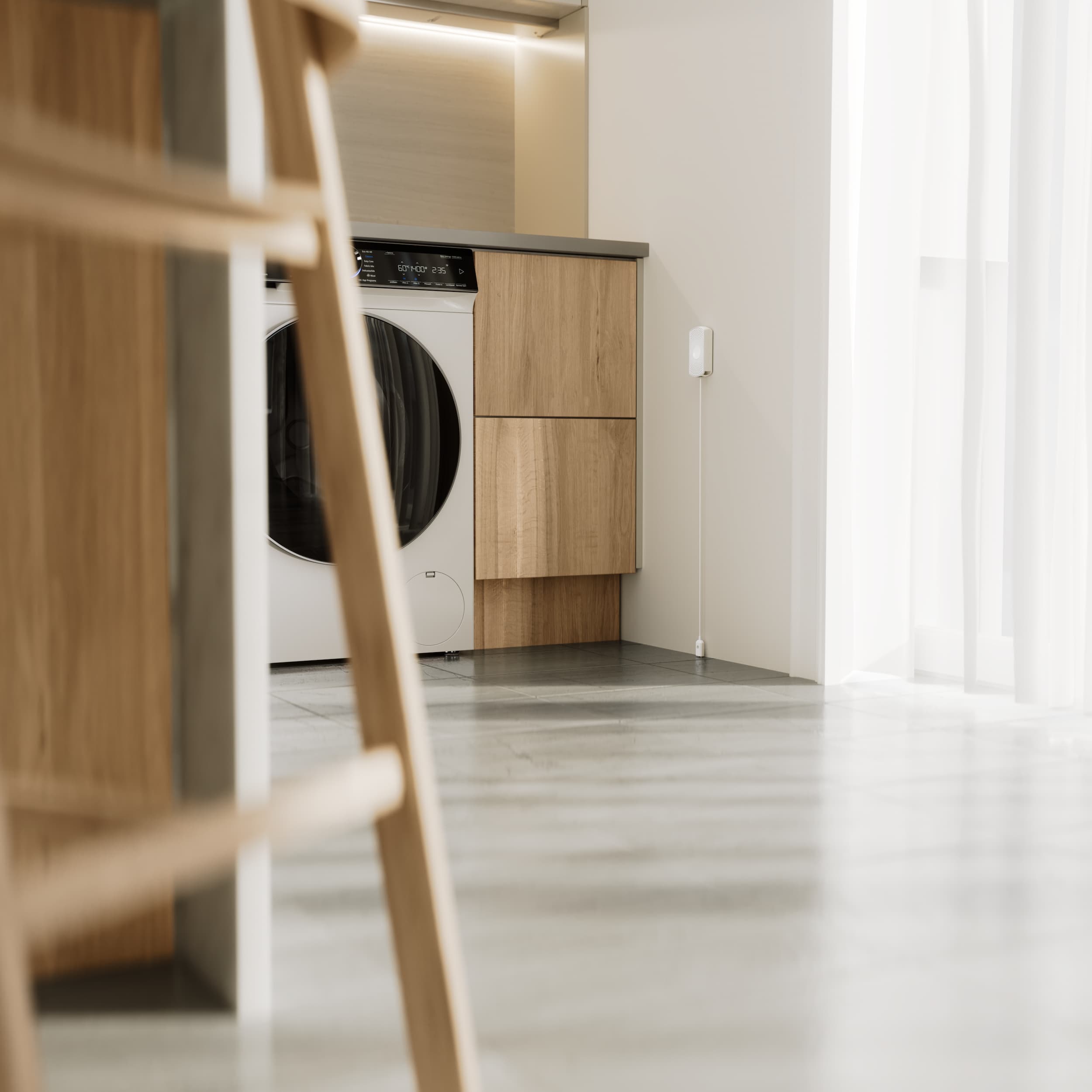 Modern laundry area with a white front-loading washing machine built into wooden cabinetry and sunlight streaming through sheer curtains.