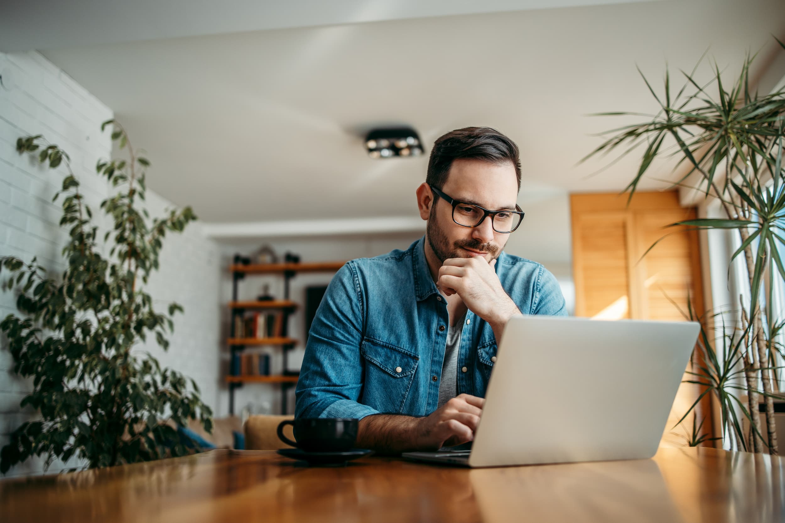 Man wearing glasses and a denim shirt working thoughtfully on a laptop at a wooden table with a black coffee cup nearby.