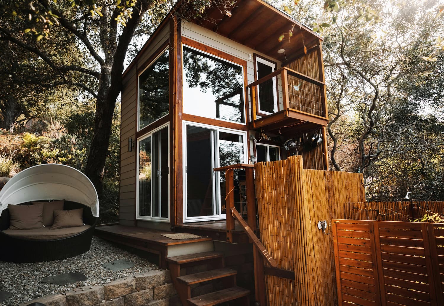 Modern tiny house with large windows, wooden balcony, bamboo privacy fence, and outdoor seating in a wooded area.