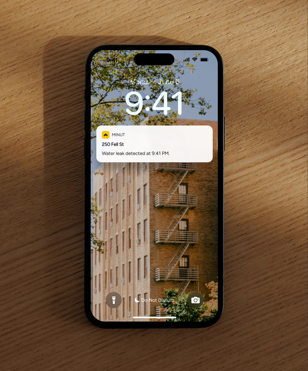Smartphone on wooden surface showing lock screen with building wallpaper and a notification of water leak detected at 9:41 PM from Minut for 250 Fell St.