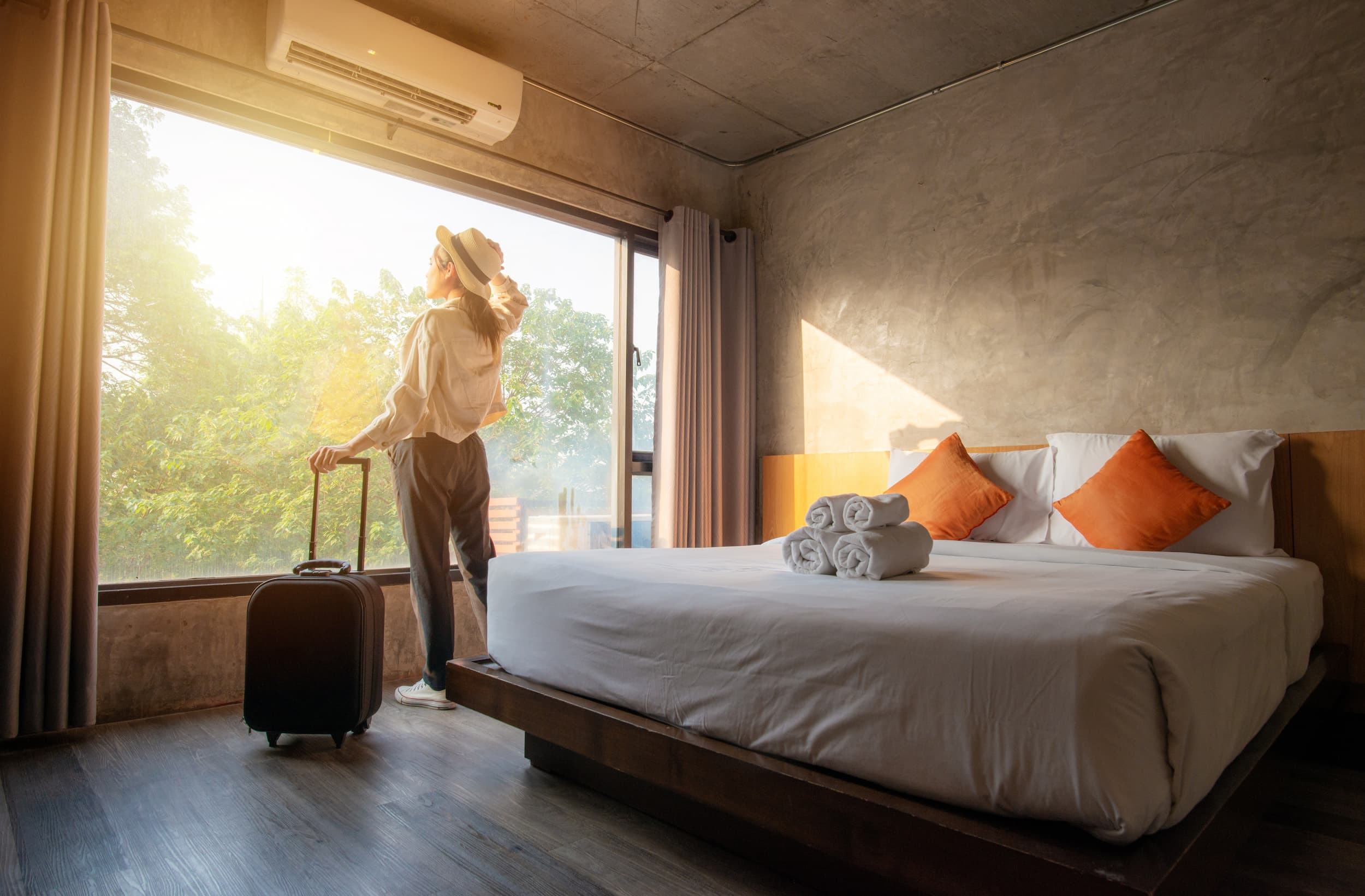 Woman with a suitcase standing by a large window in a cozy hotel room with a neatly made bed and folded towels.