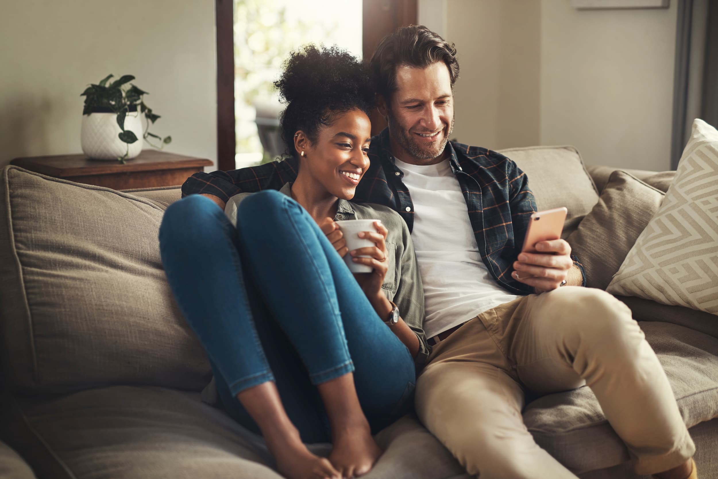 Smiling couple sitting close on a beige sofa, looking at a smartphone while the woman holds a white mug.