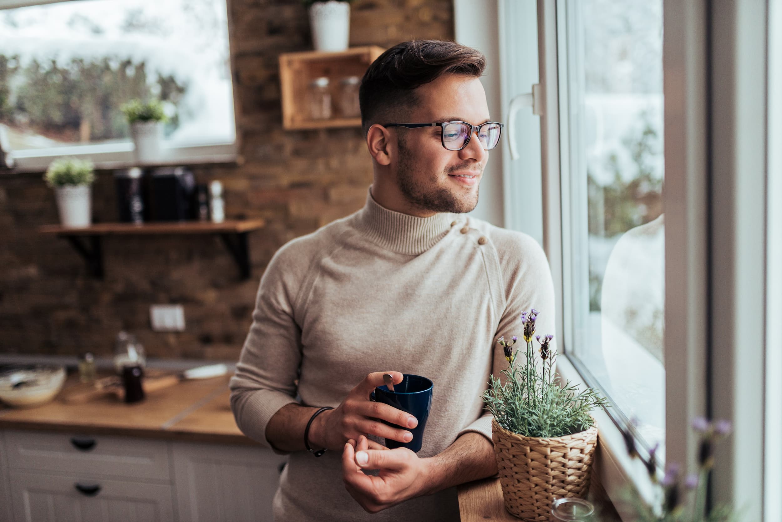 Man wearing glasses and a beige sweater holding a blue mug while looking out a window in a cozy kitchen.