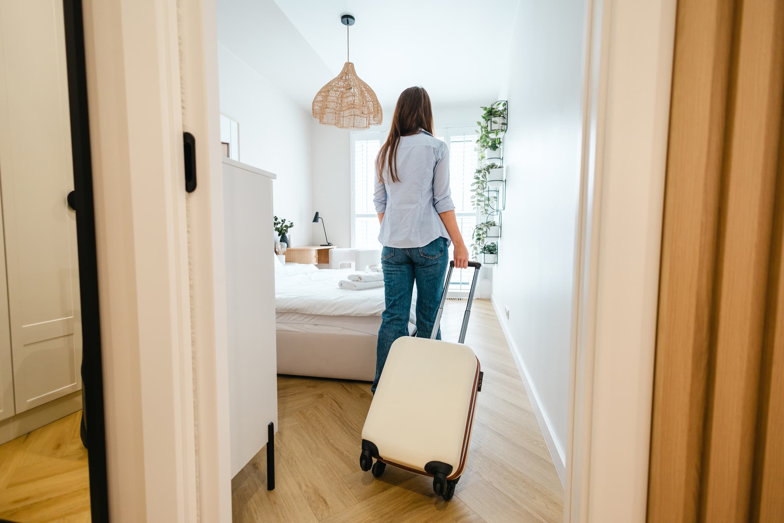 Woman with long hair pulling a beige suitcase into a bright, modern bedroom with wooden flooring and white walls.