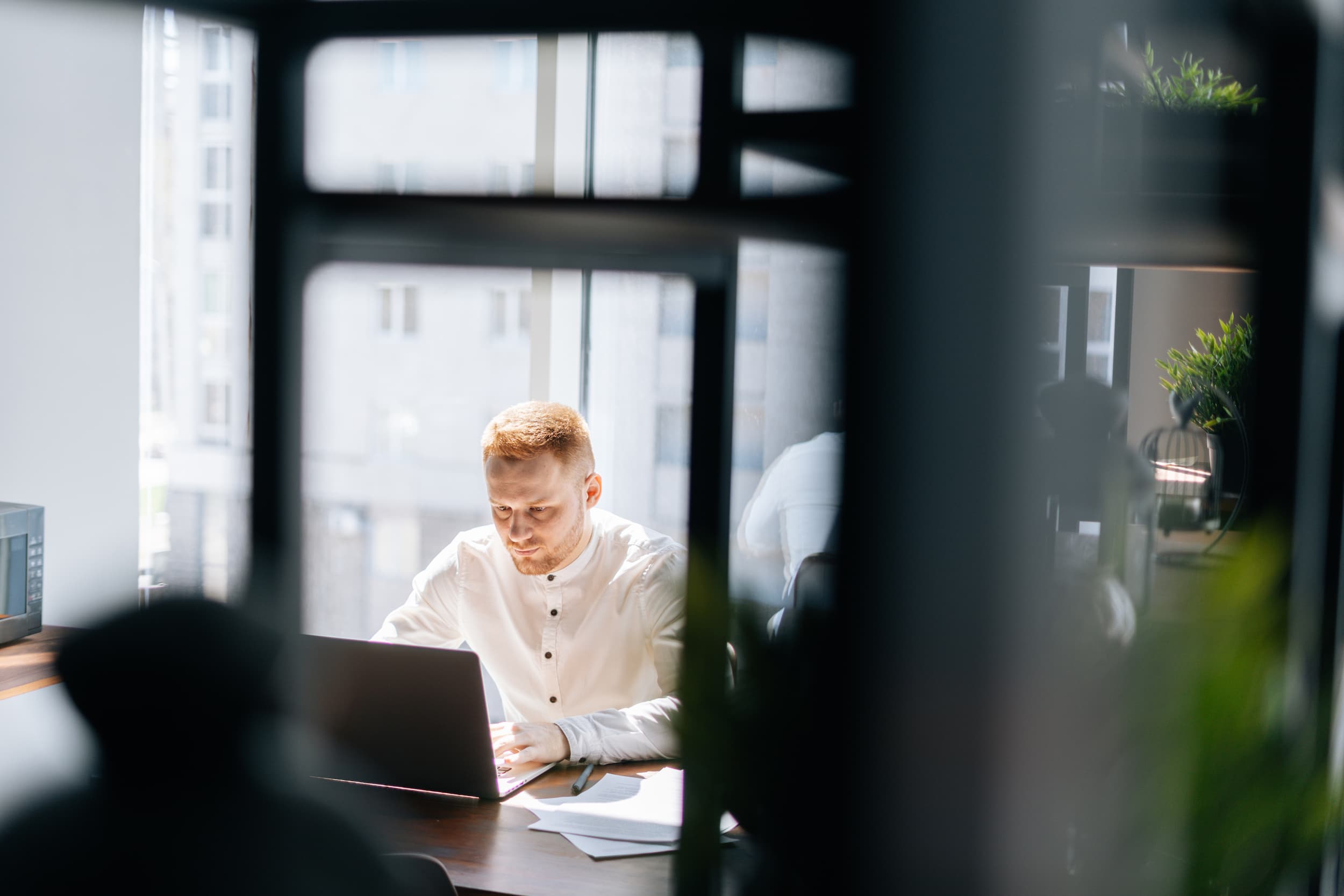 Man in a white shirt working on a laptop at a desk near a window in a bright office setting.