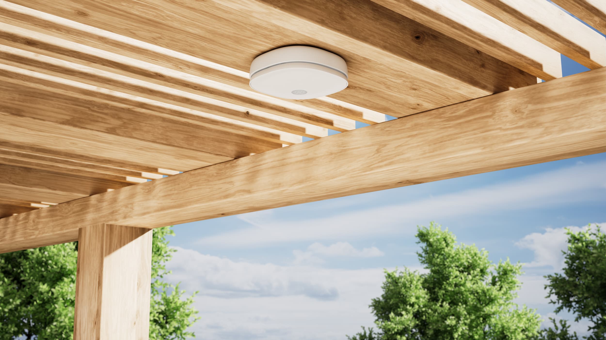 White smoke detector mounted on a wooden pergola ceiling outdoors with blue sky and green trees in the background.