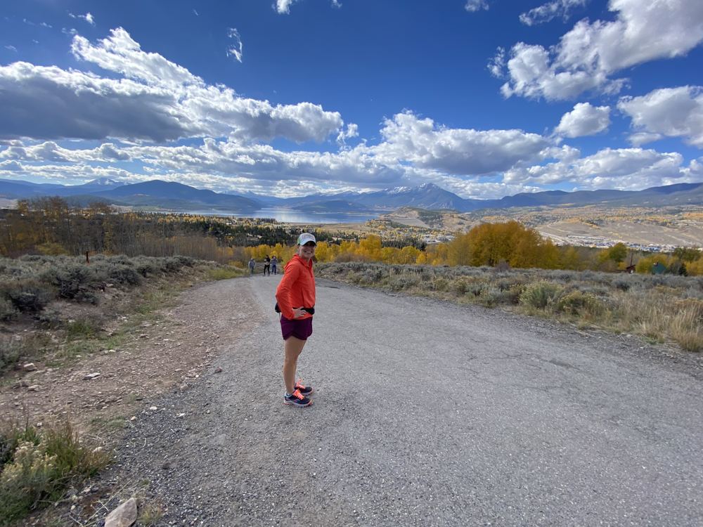 Runner in an orange jacket on a gravel road with fall foliage and mountains behind