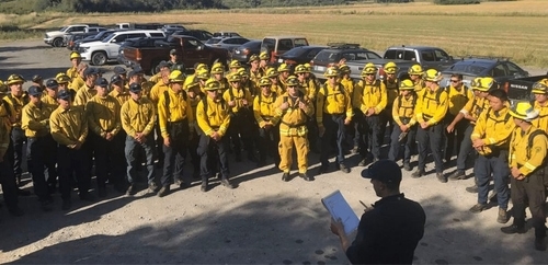 A group of firefighters in yellow uniforms gathered outdoors for a briefing, with vehicles and open fields in the background.