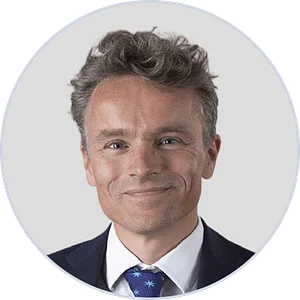 A smiling man with wavy hair, wearing a suit and tie, is pictured against a plain background in a circular frame.