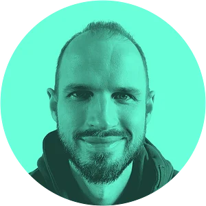 A close-up portrait of a man with short hair and a beard, smiling against a bright teal background in a circular crop.