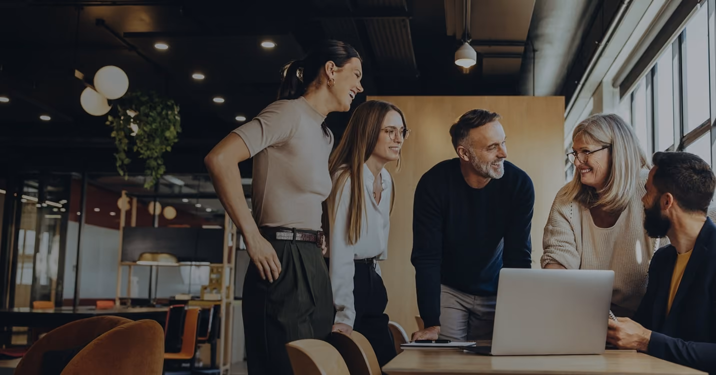 A diverse group of professionals standing around a computer, collaborating and discussing digital coaching solutions with Sharpist.