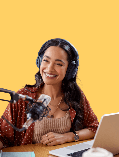Smiling woman wearing headphones speaking into a microphone while sitting at a desk with a laptop.