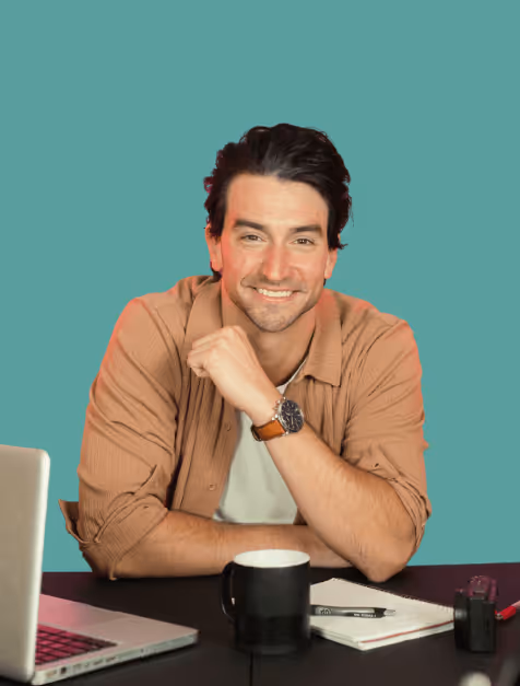 Smiling man with dark hair wearing a brown shirt sitting at a table with a laptop, black coffee mug, notebook, and pen.