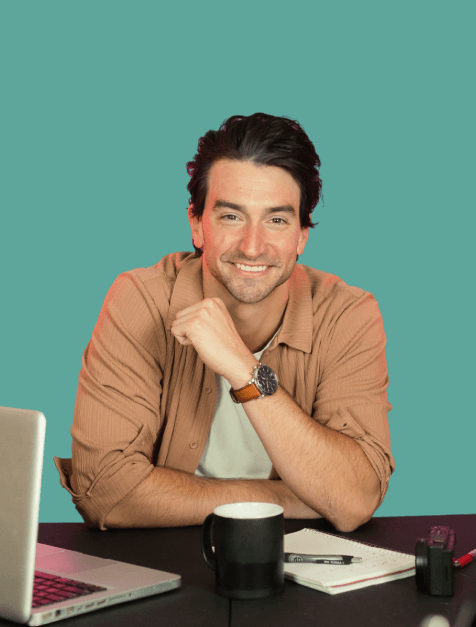 Smiling man with dark hair wearing a brown shirt sitting at a table with a laptop, black coffee mug, notebook, and pen.