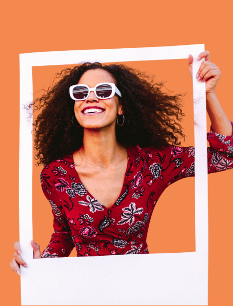 Smiling woman with curly hair wearing white sunglasses and a red floral dress holding a white photo frame against an orange background.