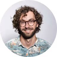 Smiling man with curly hair, glasses, and a patterned shirt against a light background.