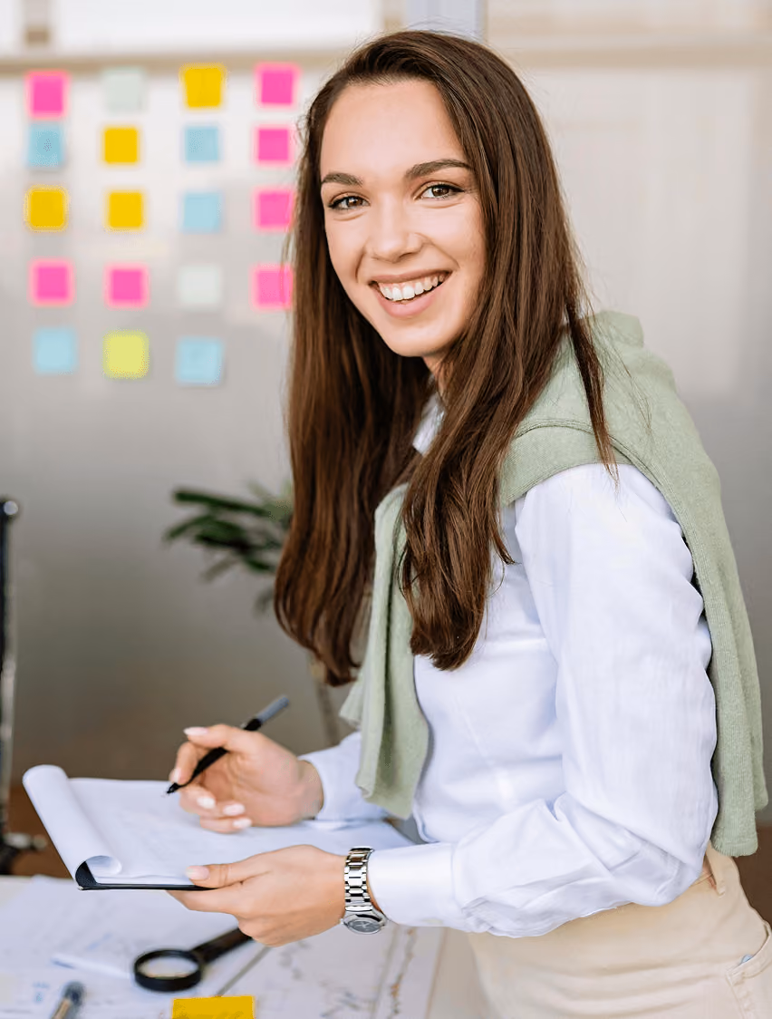 Smiling woman holding a clipboard and pen, standing in front of a wall with colorful sticky notes.