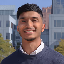 Young man with short dark hair smiling outdoors in front of office buildings and trees.