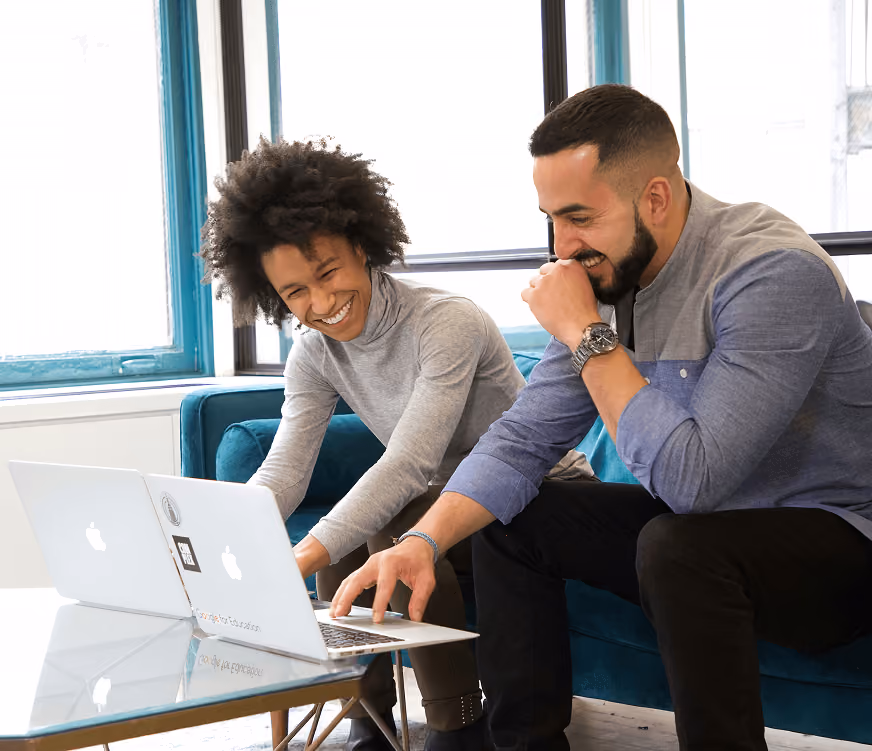 Two people sitting on a couch smiling and working on their Apple laptops side by side.