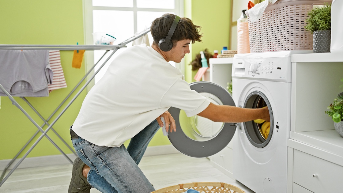 teenager wearing earbuds, white t-shirt and jeans, loading a laundry machine with clothing in a brightly lite room
