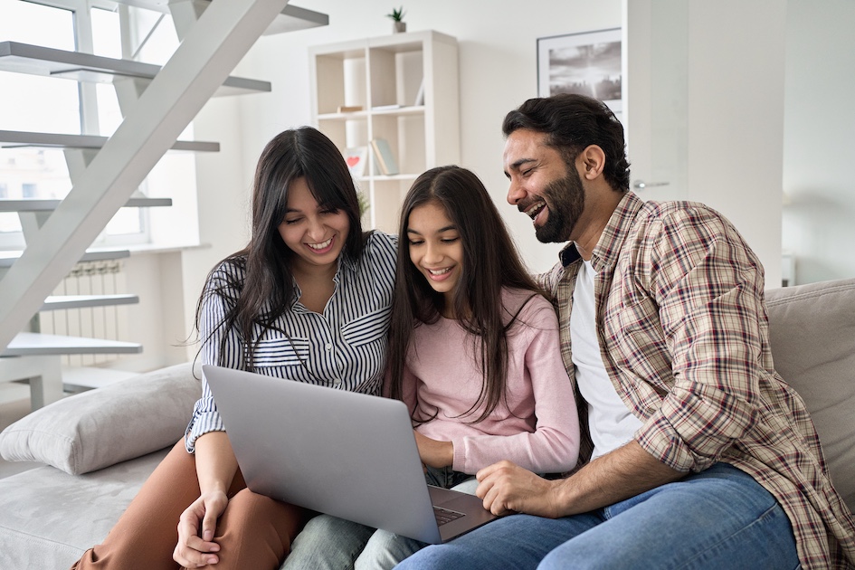 smiling mother and daughter, and father laughing, sitting on couch looking at computer