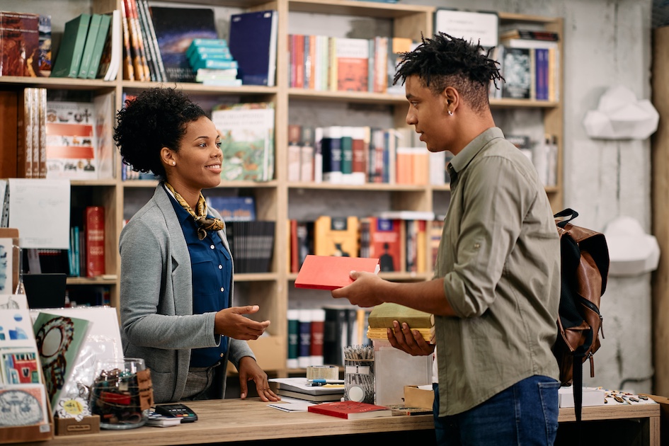 black college aged woman smiling behind counter selling book to tall black college aged man