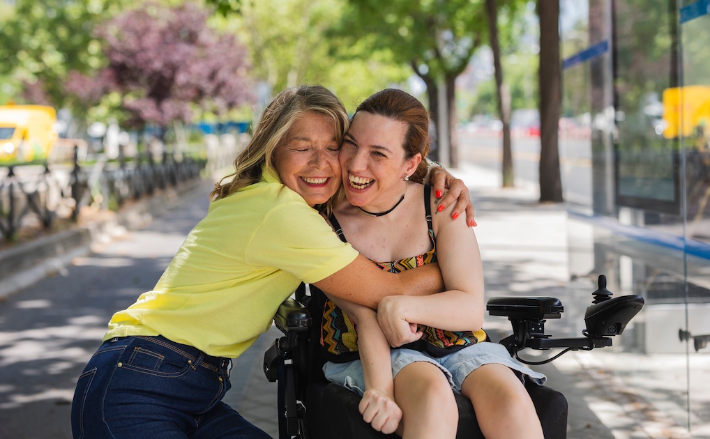 smiling mother hugging her smiling daughter who has cerebral palsy and sits in a wheelchair