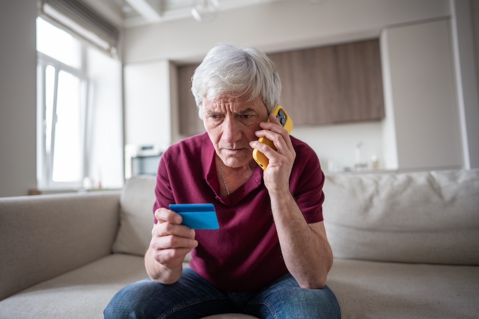 older man with white hair holding yellow phone and looking at a credit card with worried expression