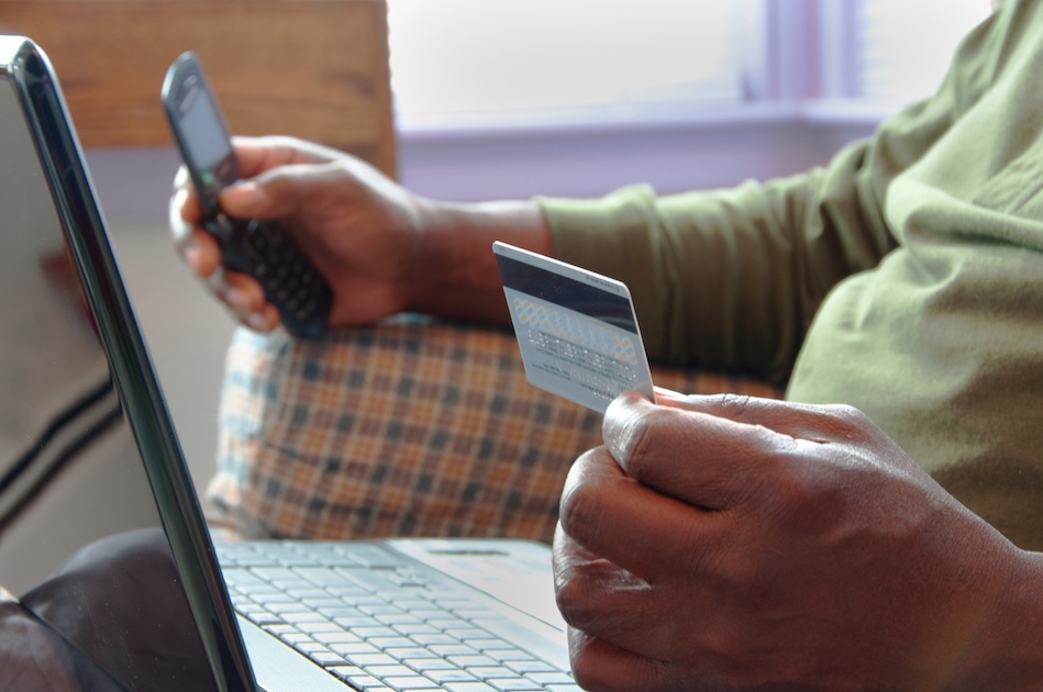 man holding credit card and cell phone near a computer appearing to be making a purchase