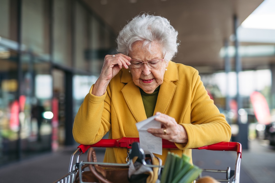 elderly white woman with white hair at a shopping cart reading a receipt wearing glasses