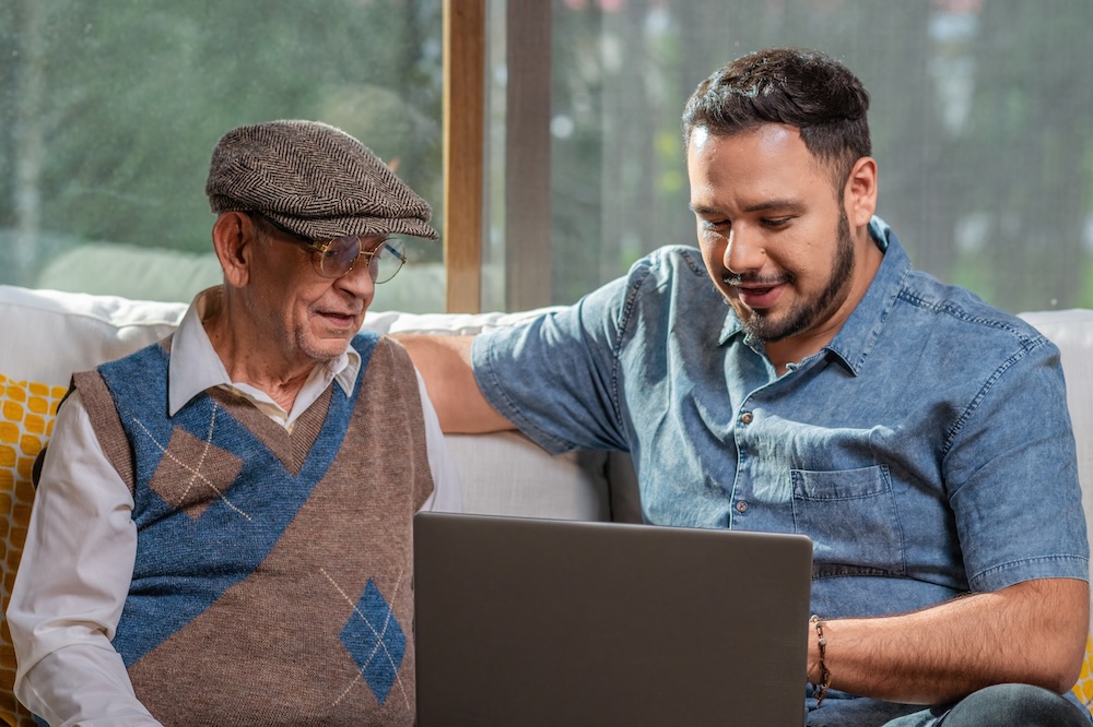 son talking to his grandfather sitting down while both looking at a laptop
