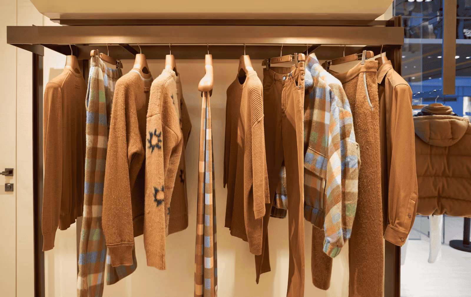 A retail display of men's clothing on a rack, featuring sweaters, pants, and a plaid jacket with a color palette of brown, tan, and blue.