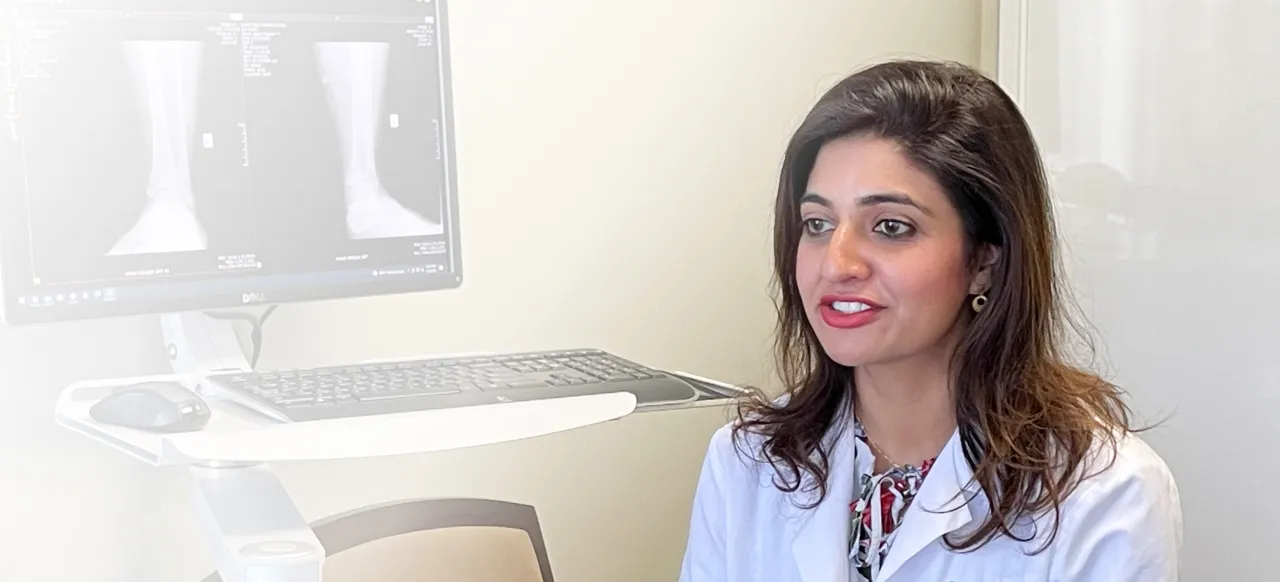 Female doctor in white coat speaking, with X-ray images on a computer screen in the background.