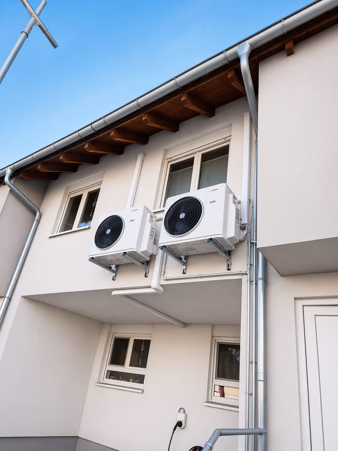 Außenwand eines modernen Hauses mit zwei angebrachten Bosch-Klimageräten unter einem braunen Holzdach und einem klaren blauen Himmel.