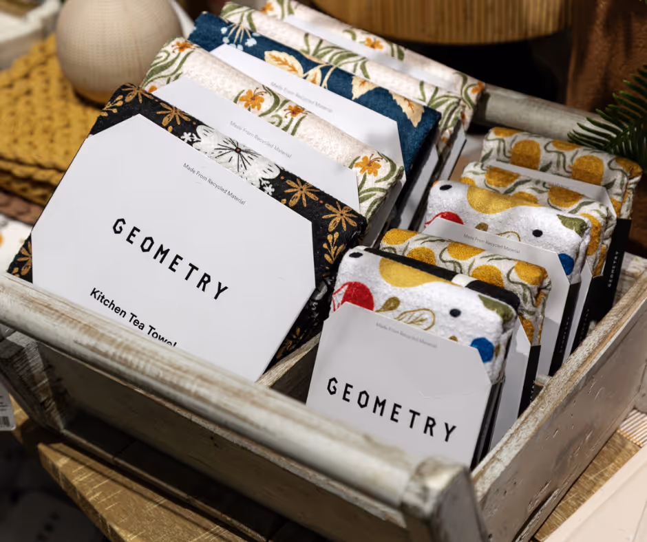 Wooden tray holding assorted packaged kitchen tea towels and dishcloths with floral and geometric patterns labeled 'GEOMETRY'.