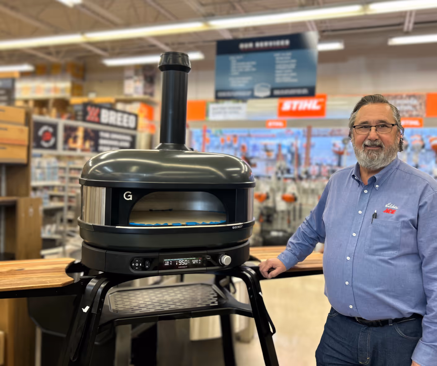 Man wearing glasses and a blue shirt stands next to a black Gozney pizza oven on a stand in a store.