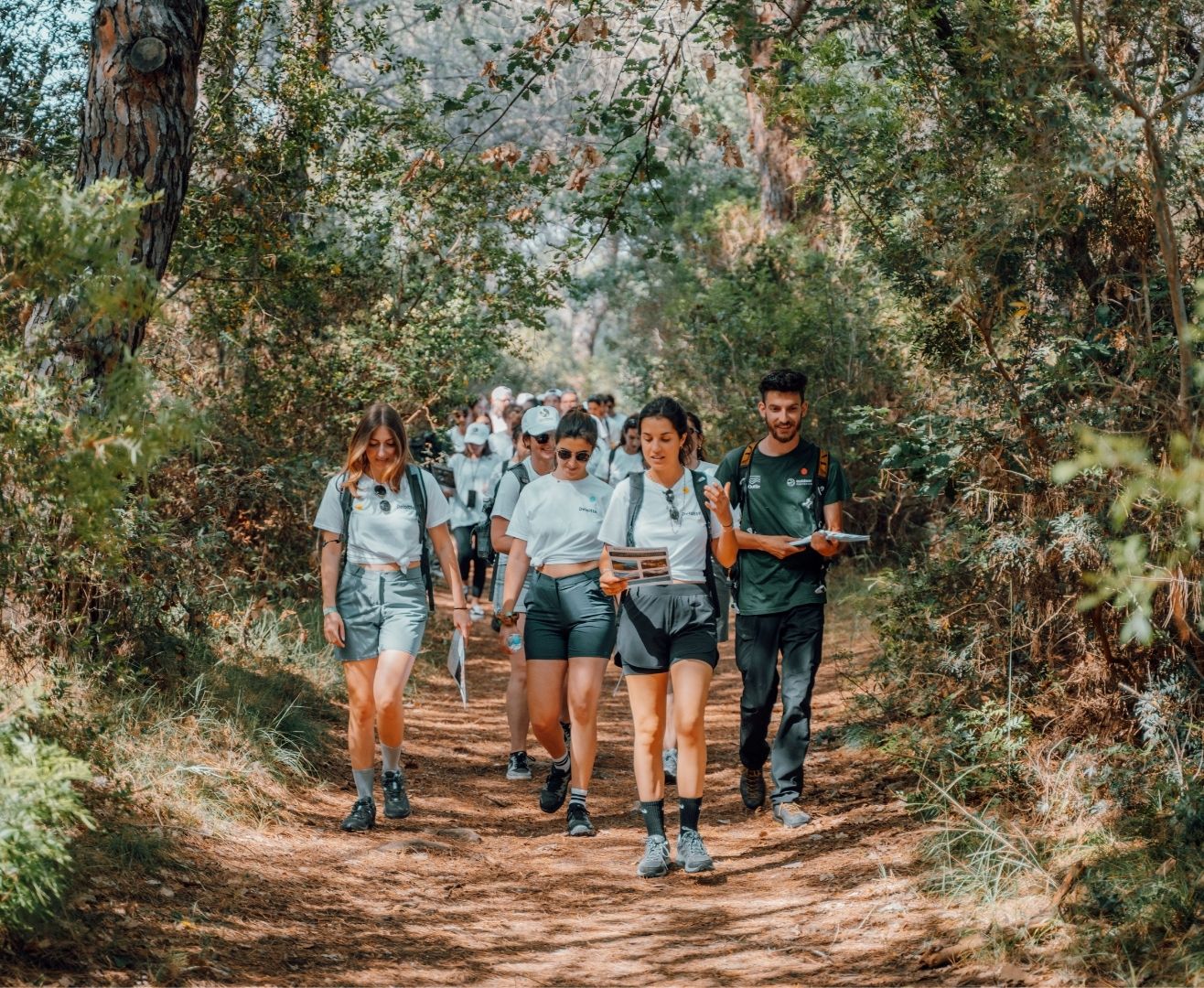 Gruppo di giovani in escursione su un sentiero di bosco, alcuni leggono mappe e altri camminano insieme.