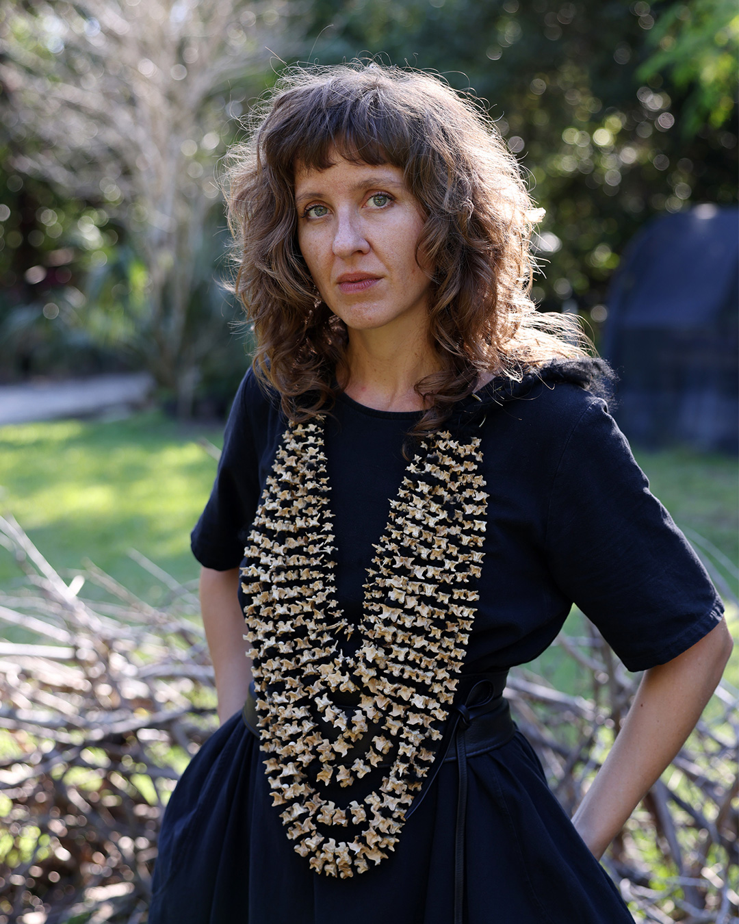 Rebecca inside an outdoor installation of her work, wearing black dress and a garland of python bones