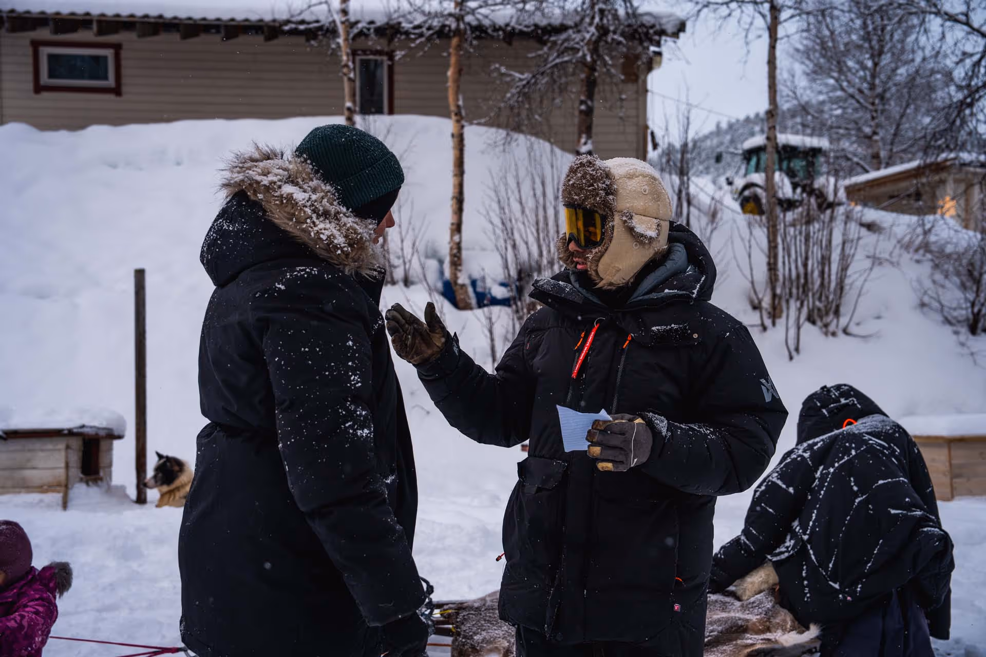 Two people dressed in heavy winter clothing talking in a snowy outdoor setting with a dog and wooden doghouses nearby.
