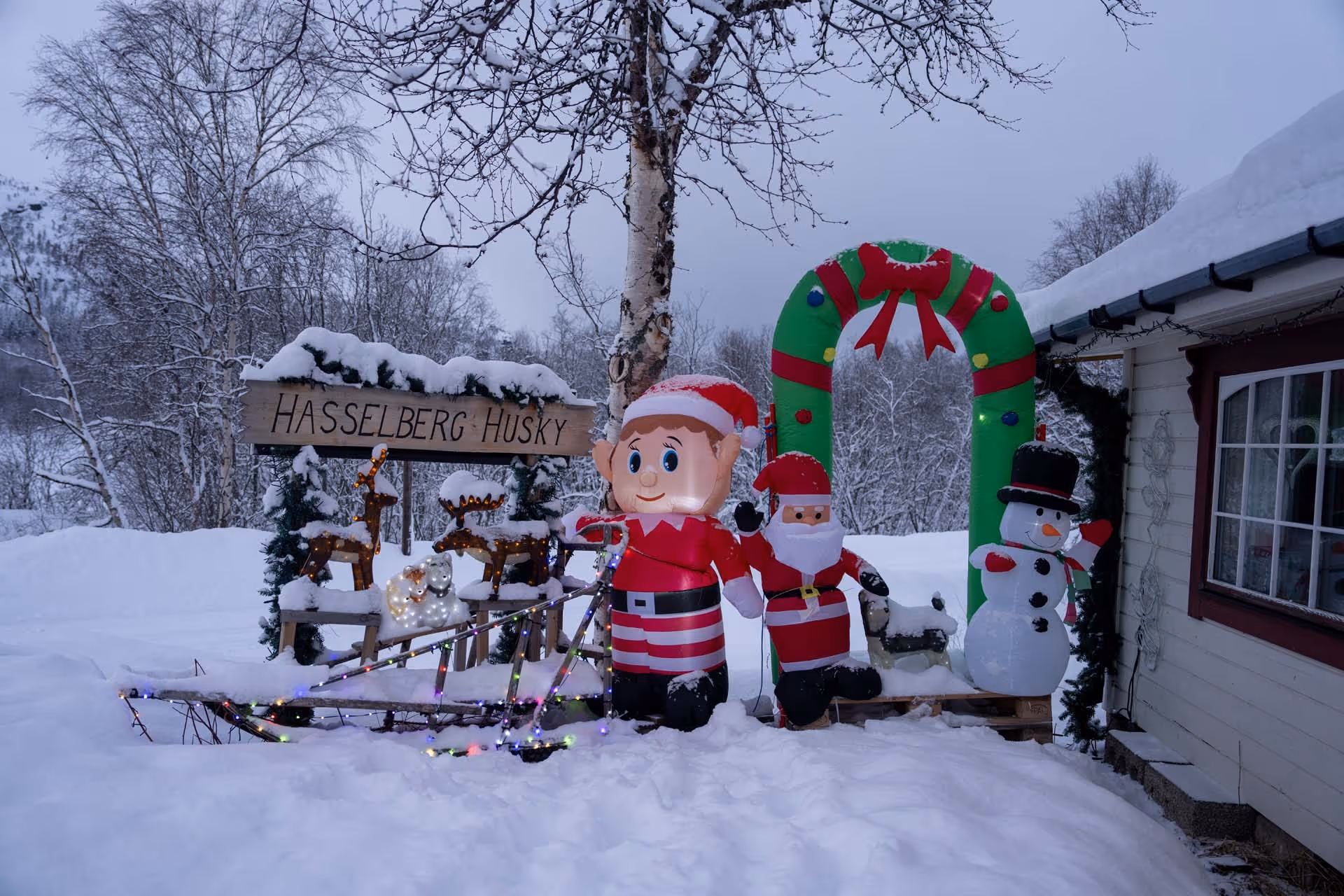 Snow-covered outdoor Christmas display with inflatable Santa, elf, snowman, reindeer figures, and a sign reading Hasselberg Husky.