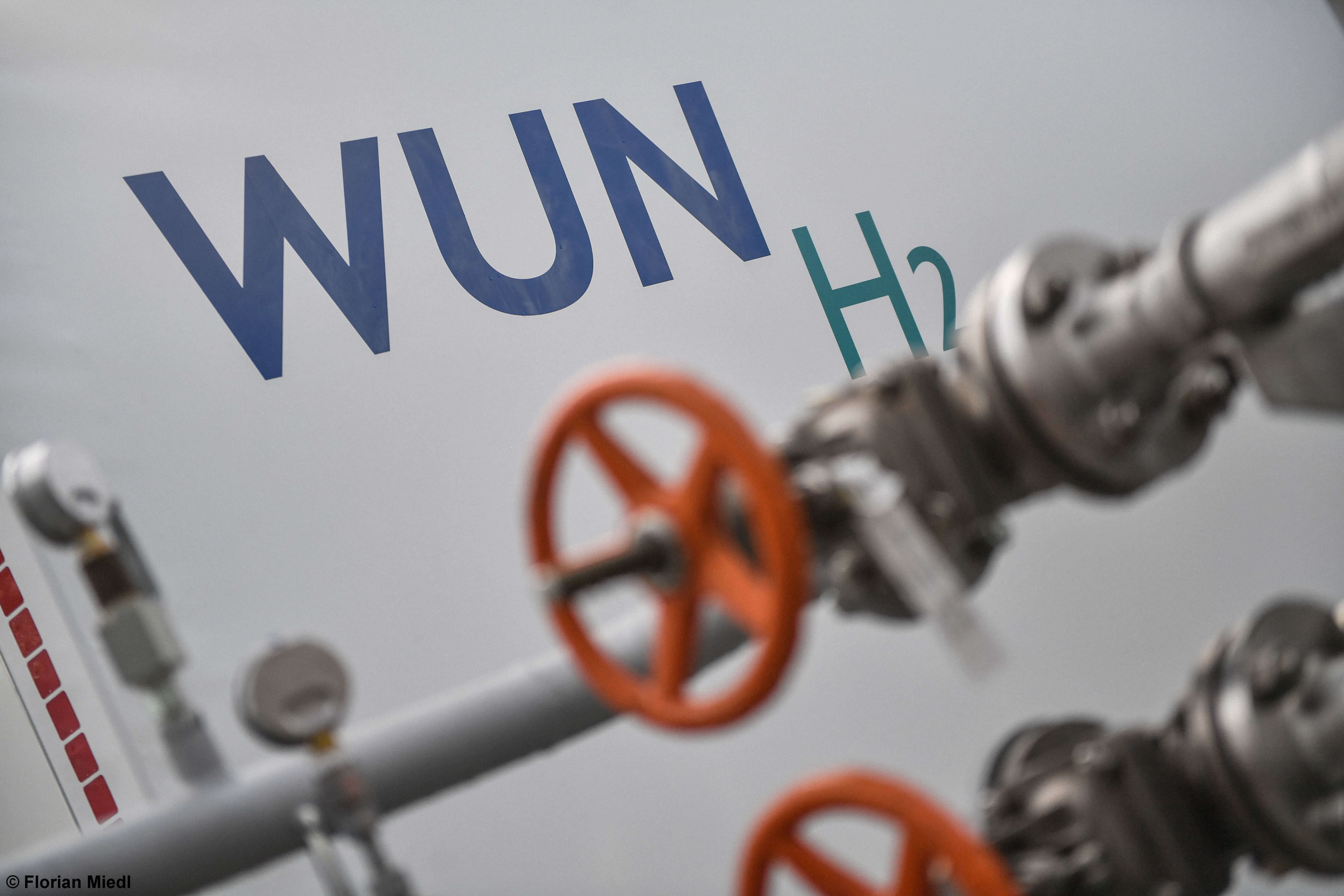Detailaufnahme eines industriellen Rohrleitungssystems mit orangefarbenen Handrädern im Vordergrund und dem Schriftzug „WUN H₂“ auf einem großen weißen Tank im Hintergrund – symbolisiert die Infrastruktur zur Erzeugung oder Speicherung von blauem Wasserstoff.